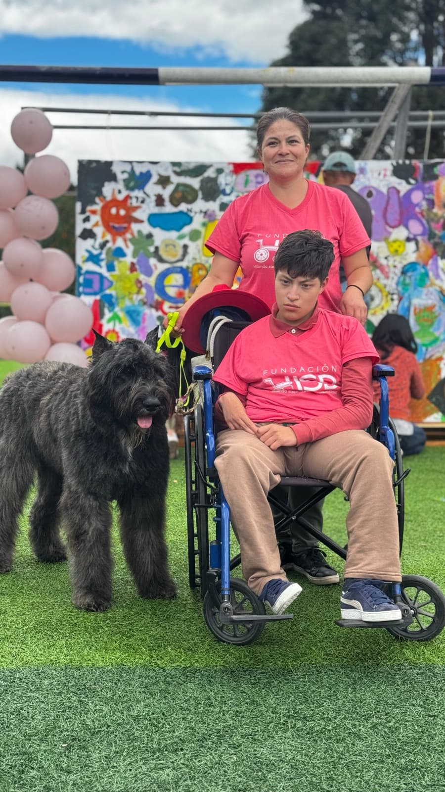 Beneficiario de HSD en silla de ruedas, con camiseta de la fundación, acompañado de un voluntario y un perro en un evento